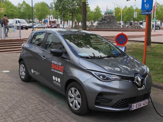 Grijze elektrische deelauto van Mobilize Share geparkeerd en opgeladen in een stedelijke omgeving. (vergroot foto)